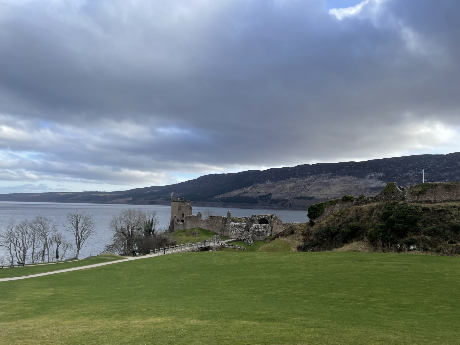 A wide view of Urquhart Castle ruins on a grassy hill beside Loch Ness, with mountains in the distance under a cloudy sky.