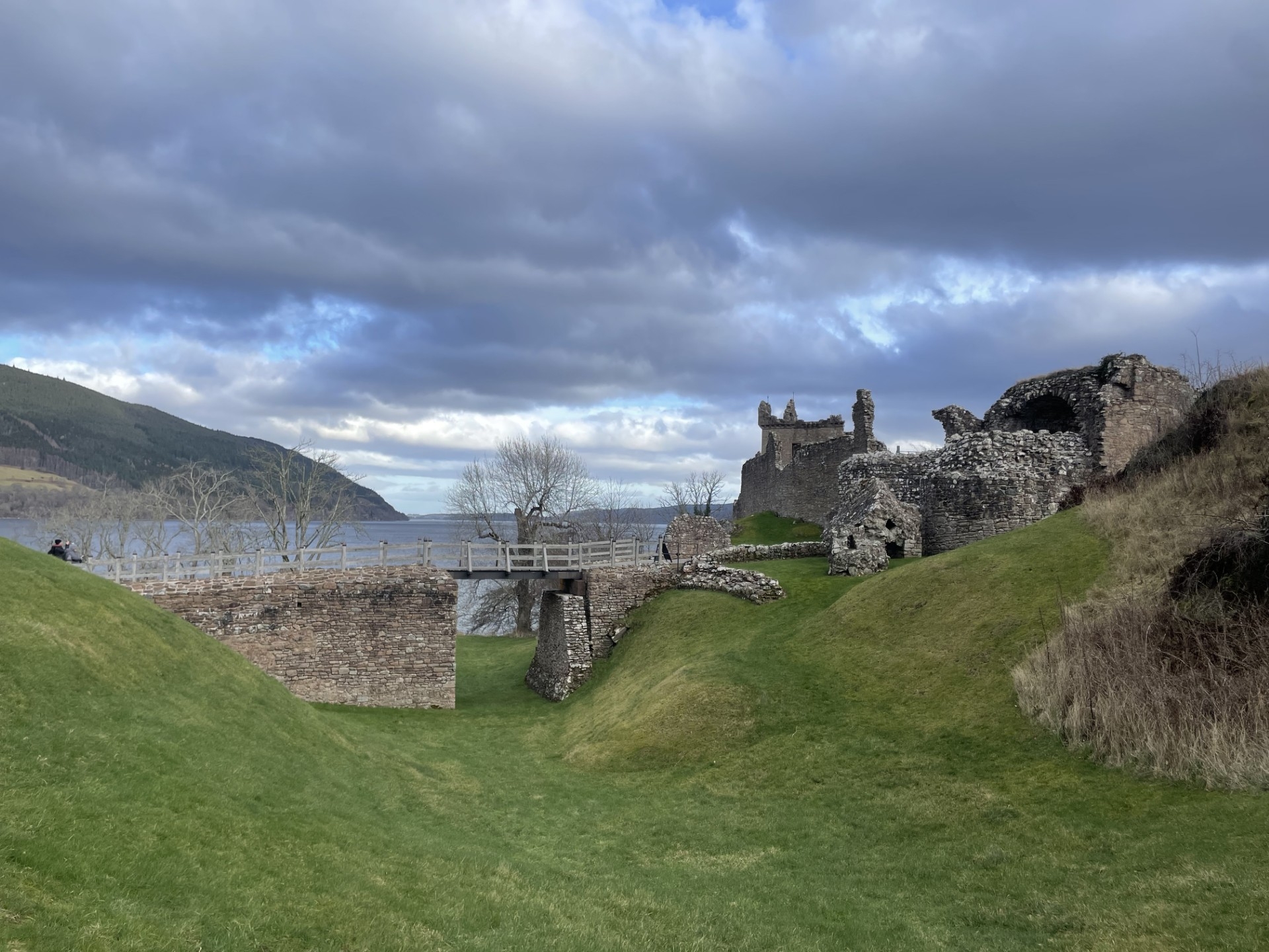 A grassy landscape leading to sections of the castle ruins, connected by a wooden footbridge spanning a dip in the land, with the loch visible in the background.
