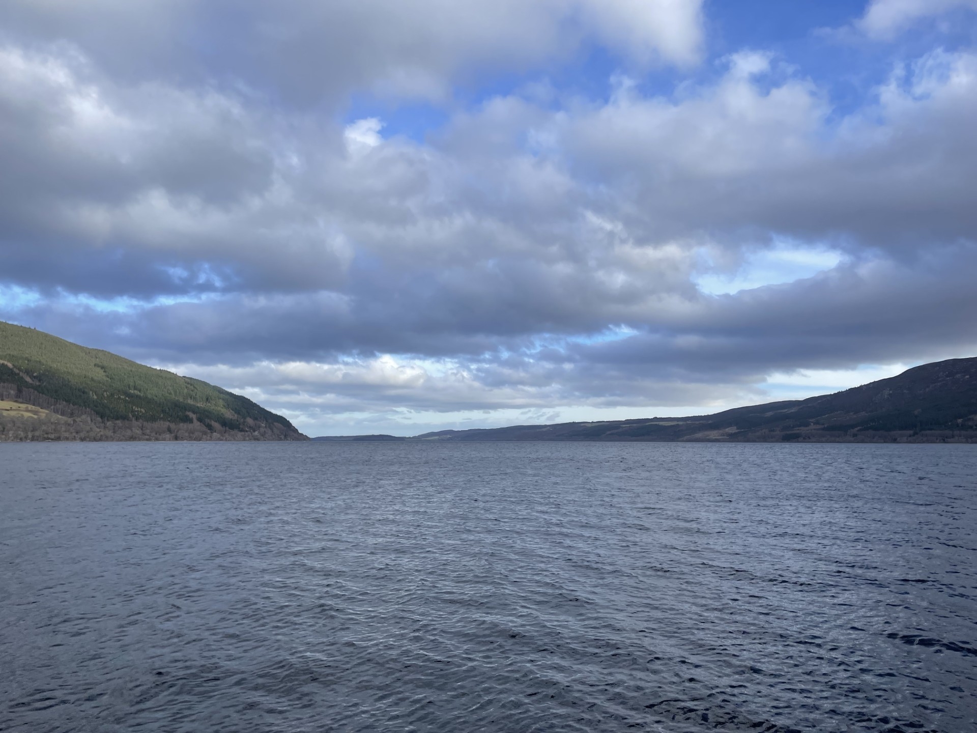 A broad, open view of Loch Ness stretching into the distance, surrounded by hills beneath a dramatic sky of clouds and patches of blue.