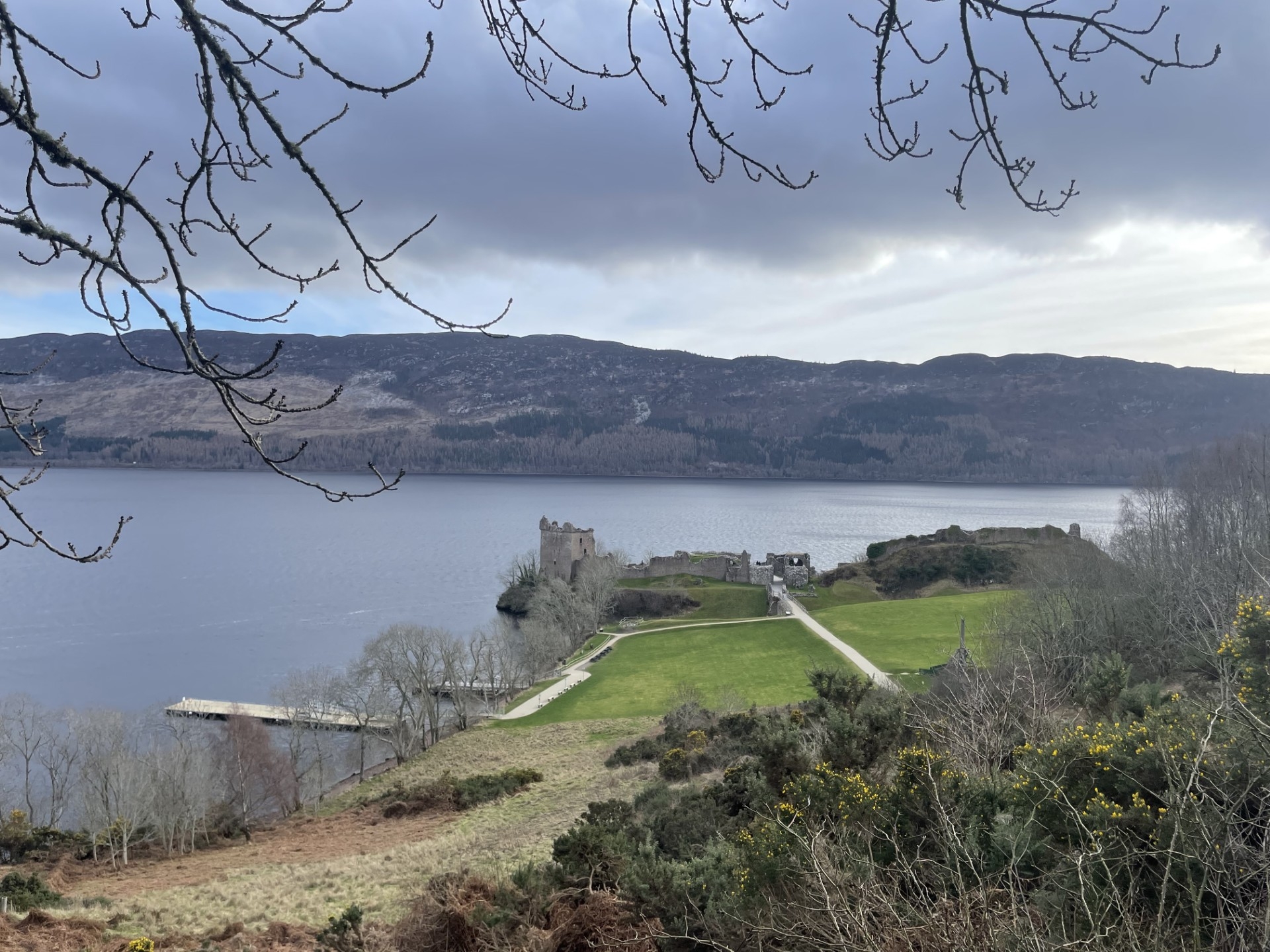 A high vantage point looking down toward Urquhart Castle by the water, framed by bare tree branches, with sweeping views of the loch and surrounding hills.