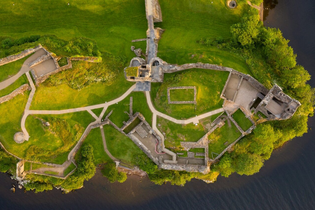 Aerial view of the ruins of Urquhart Castle sitting on a grassy promontory beside Loch Ness, showing the full layout of the remaining stone walls, courtyards, and pathways surrounded by lush greenery and the dark waters of the loch.
