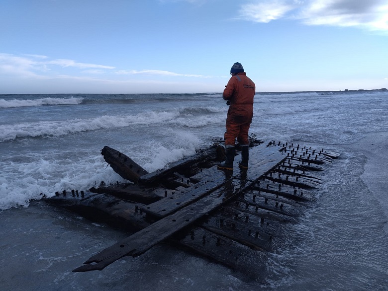 A person in waterproof gear stands on the remains of a wooden shipwreck on a beach. Waves crash around the exposed timbers as the sea stretches into the distance under a cloudy sky.