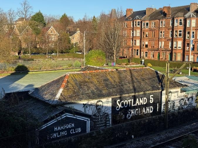View of a bowling club with park and tenement housing seen beyond it.