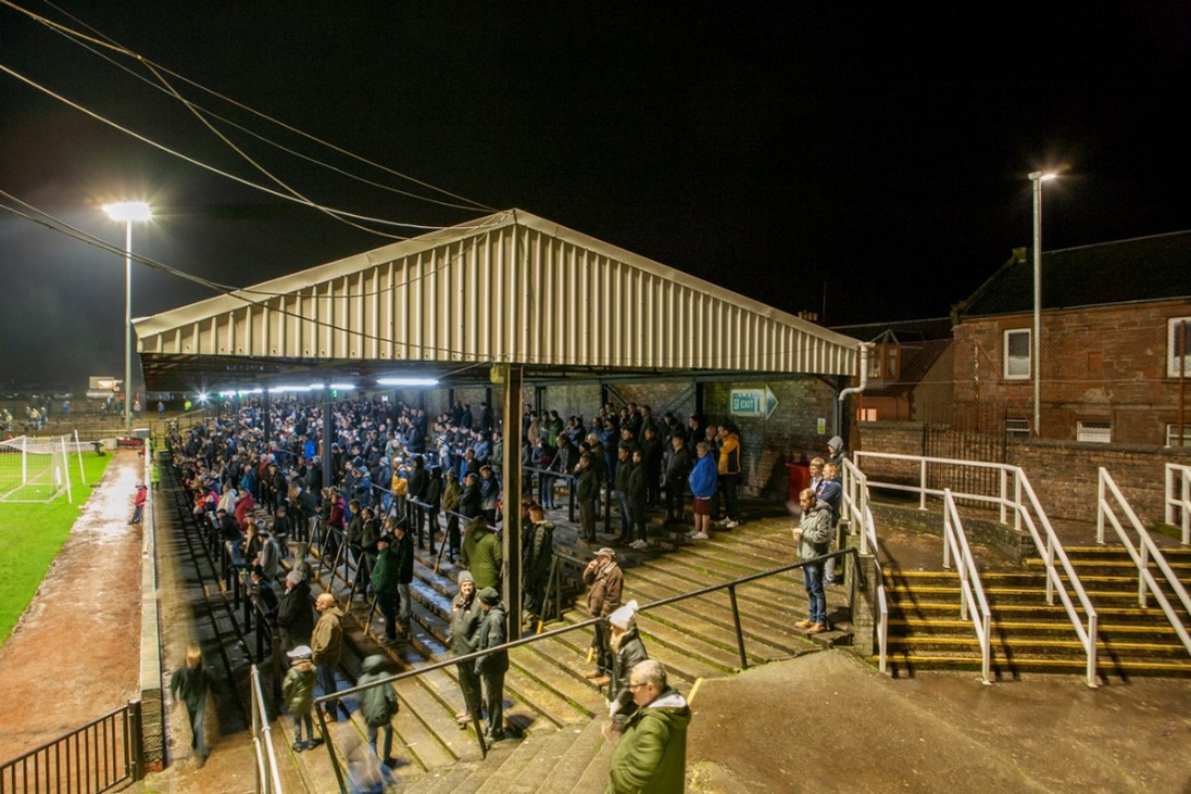 Crowd of football supporters standing on covered terracing at a floodlit football ground during a nighttime match.