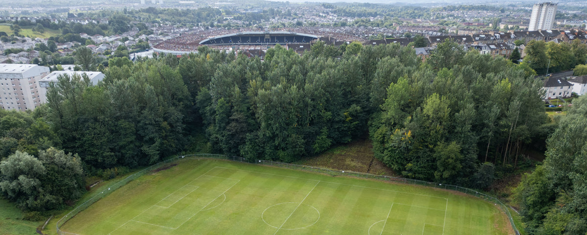 Aerial view of a football pitch bordered by dense woodland, with urban housing and a stadium visible in the background.