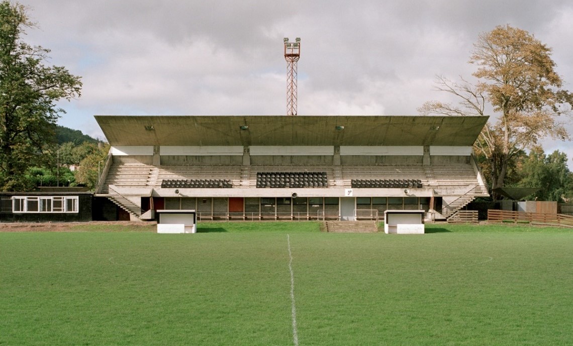 Front view of a small covered football stand facing a grass pitch, with rows of seats, floodlight tower and trees under a cloudy sky.