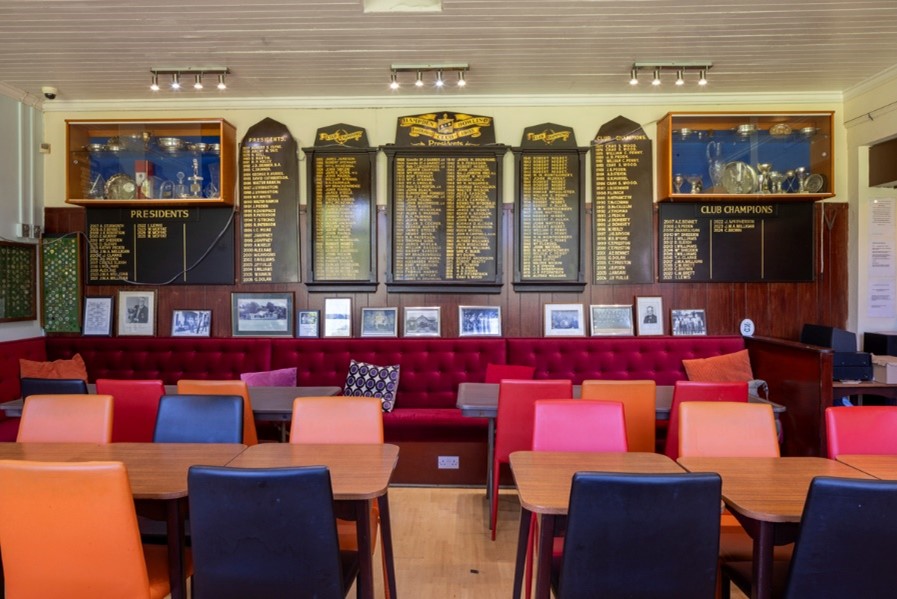 Interior of a club lounge with colourful chairs and tables, red seating, trophy cabinets and honour boards displayed on wood-panelled walls.