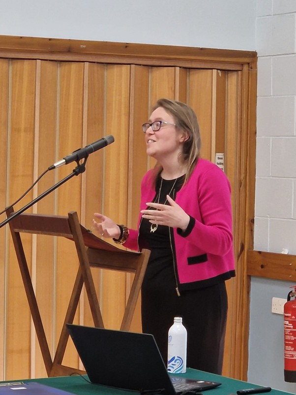 Woman in a pink jacket gives a talk into a microphone.