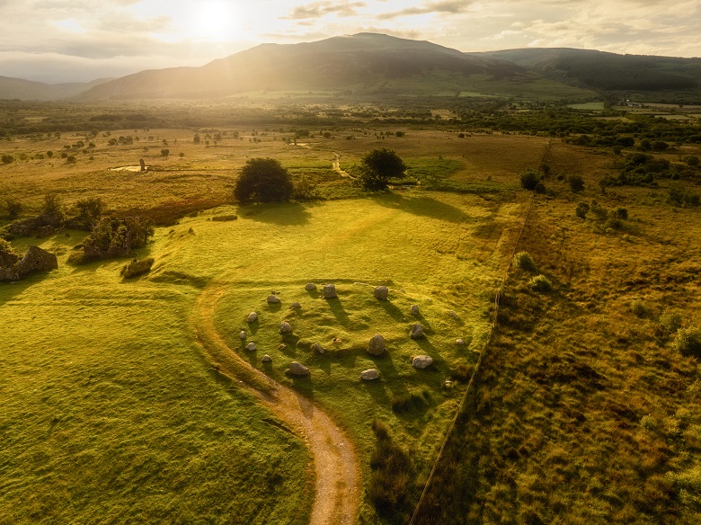Aerial view of a small stone circle set within a grassy enclosure at Machrie Moor on Arran. A winding path curves around the monument, while the wider moorland landscape stretches towards distant hills and mountains. Warm evening sunlight casts long shadows across the grass, highlighting the stones and surrounding archaeological features.