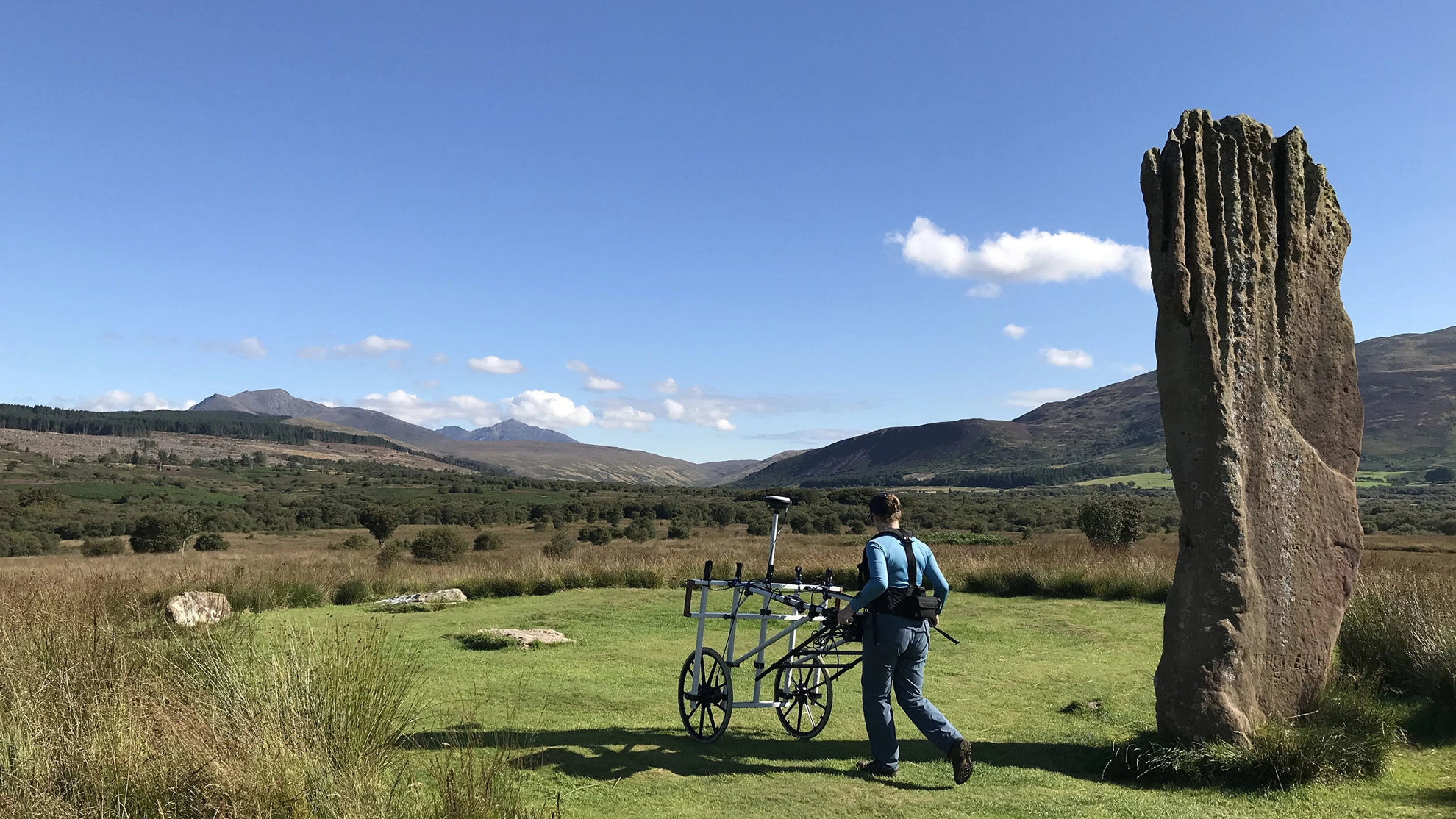 A geophysical survey in progress at Machrie Moor on Arran. A surveyor pushes a wheeled geophysical survey frame across grass beside a tall standing stone.