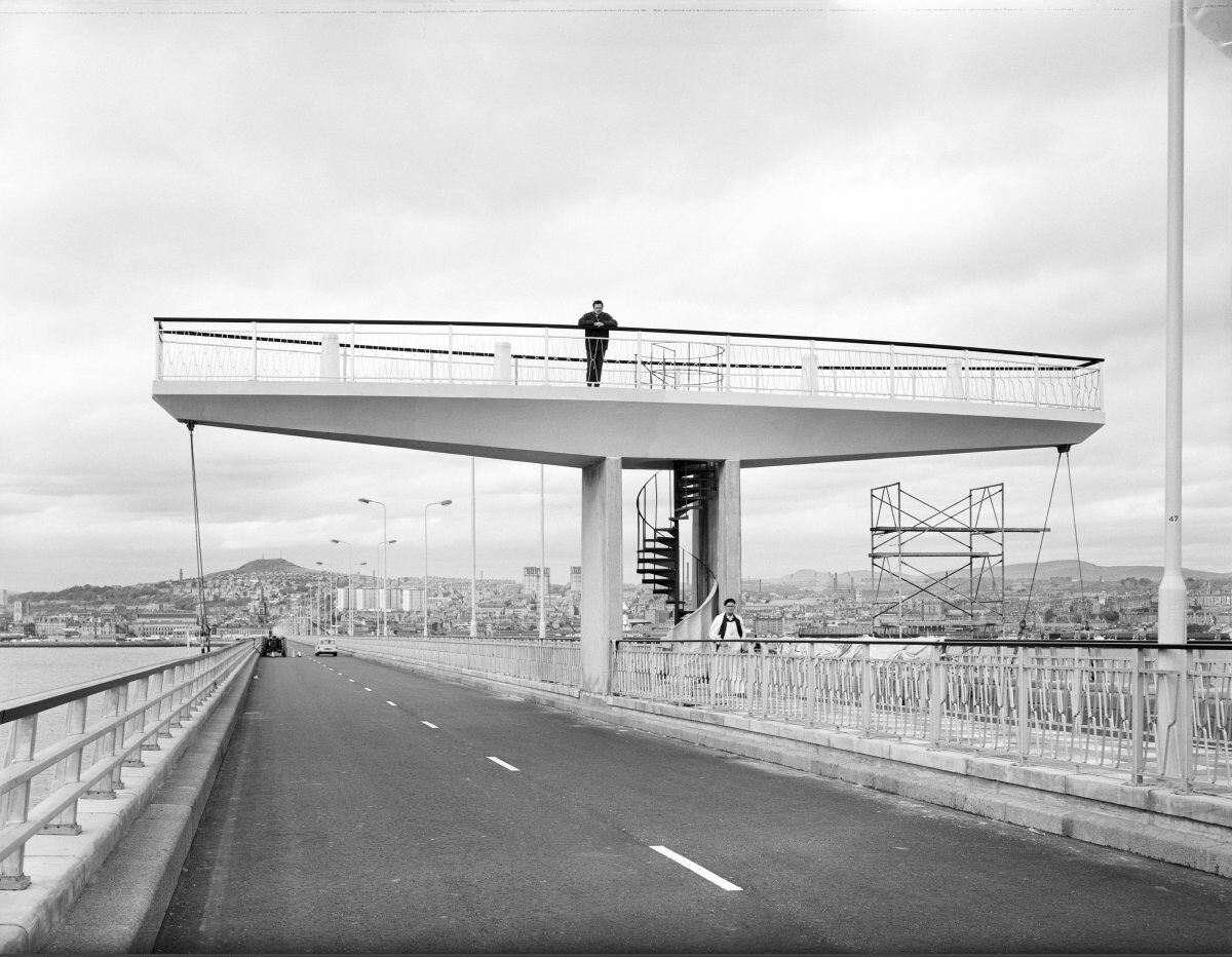 A black-and-white view looking along a bridge carriageway toward a large elevated observation platform. The platform projects over the road and is supported by concrete columns, with a spiral staircase leading up from the pedestrian walkway. A few people are visible on and beneath the structure, while the waterfront cityscape stretches across the background.