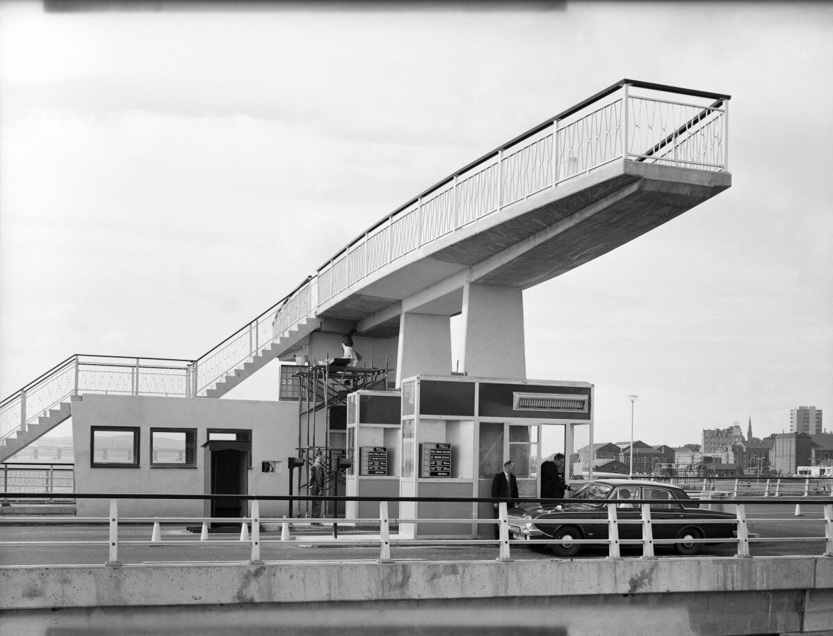 Image 11: A black-and-white photograph showing a toll booth complex on a bridge shortly before completion or opening. An elevated pedestrian bridge crosses above the toll area, connecting stairways on either side. A car and several people stand near the toll booths, with urban buildings visible beyond.
