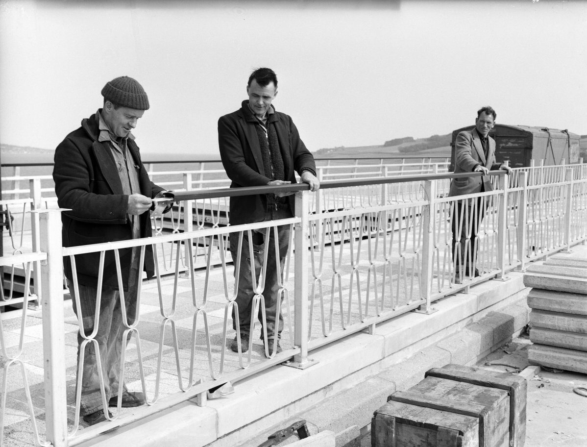 Black-and-white photograph of three workers installing decorative metal railings on a bridge walkway. Construction materials are stacked nearby, with water and shoreline visible beyond the bridge.