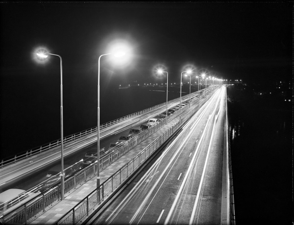 Black-and-white night photograph of a bridge illuminated by streetlights, with long-exposure light trails from moving vehicles stretching along both carriageways.
