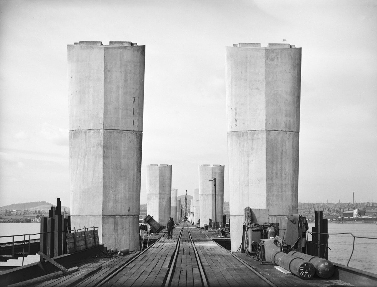 Black-and-white photograph showing a bridge under construction. Tall concrete support piers rise from a timber work platform with rails running down the centre, while construction equipment and a worker are visible along the deck.