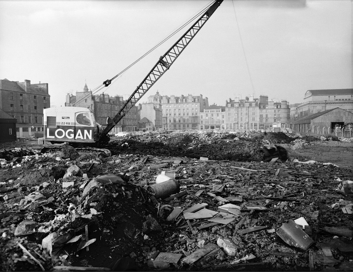 Black-and-white photograph of a large building site covered in rubble and debris, with a crawler crane marked “LOGAN” standing in the foreground. Rows of multi-storey city buildings and parked cars are visible in the background.