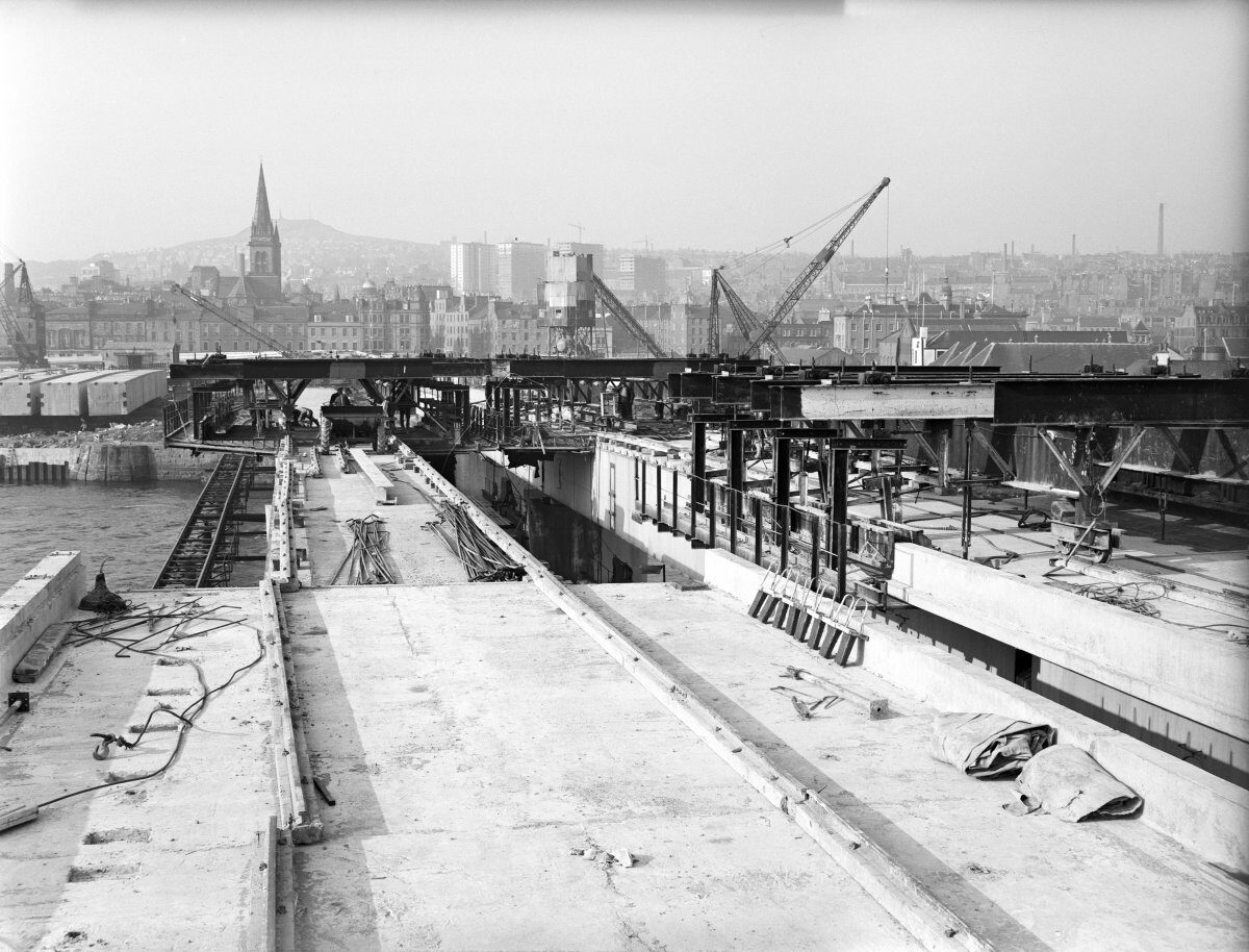 Black-and-white photograph of a harbour-side construction area with water in the foreground, piles of rubble along the shoreline, and large warehouse buildings behind. Historic city buildings, church spires and cranes rise in the background.
