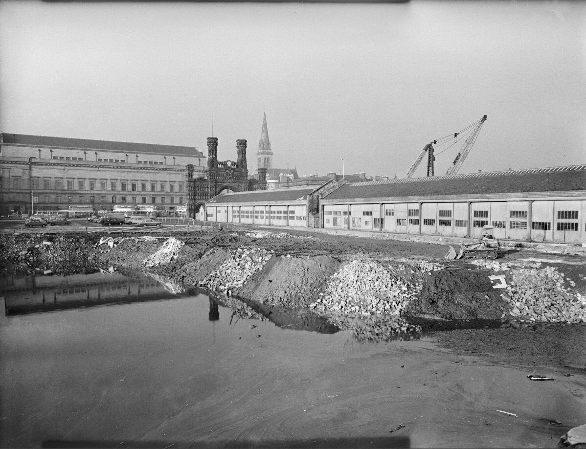 Black-and-white photograph of a harbour basin bordered by a cobbled quayside. Warehouses and dockside cranes stand across the water, with industrial buildings lining the waterfront.