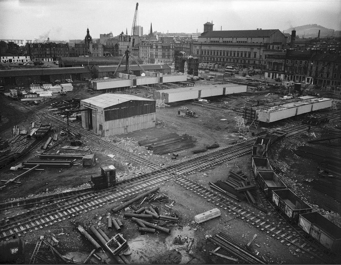 Black-and-white photograph of a large construction yard crossed by multiple rail tracks. Steel beams, construction materials and temporary buildings are scattered across the site, with city buildings and cranes visible in the background.