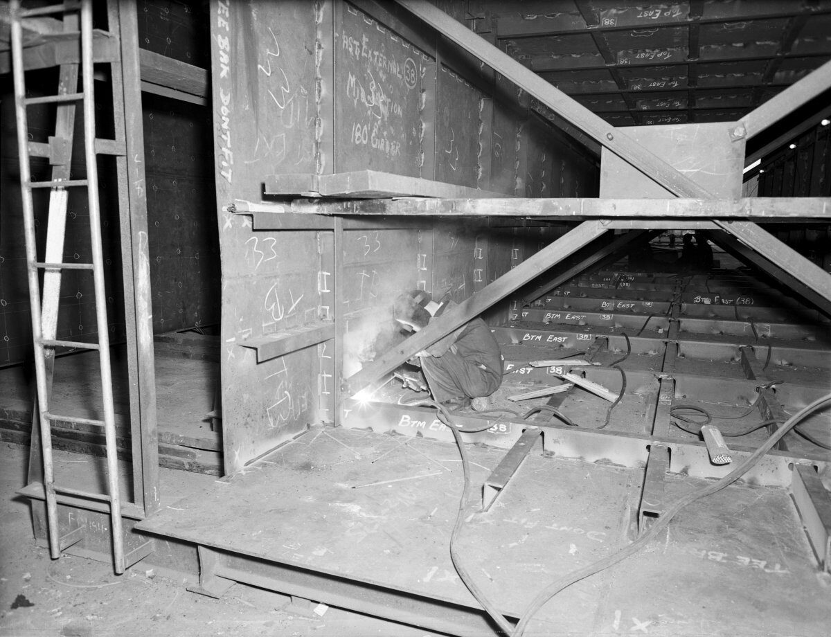 Black-and-white photograph of a worker welding inside a steel bridge structure. Large steel beams, cross-bracing, cables and a ladder surround the workspace, with sparks visible at the welding point.