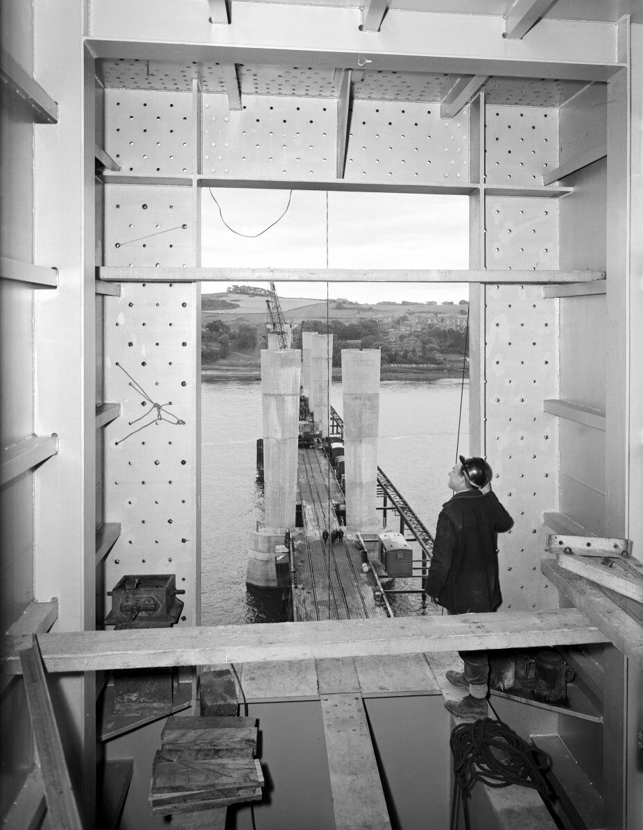 Black-and-white photograph of a worker welding inside a steel bridge structure. Large steel beams, cross-bracing, cables and a ladder surround the workspace, with sparks visible at the welding point.