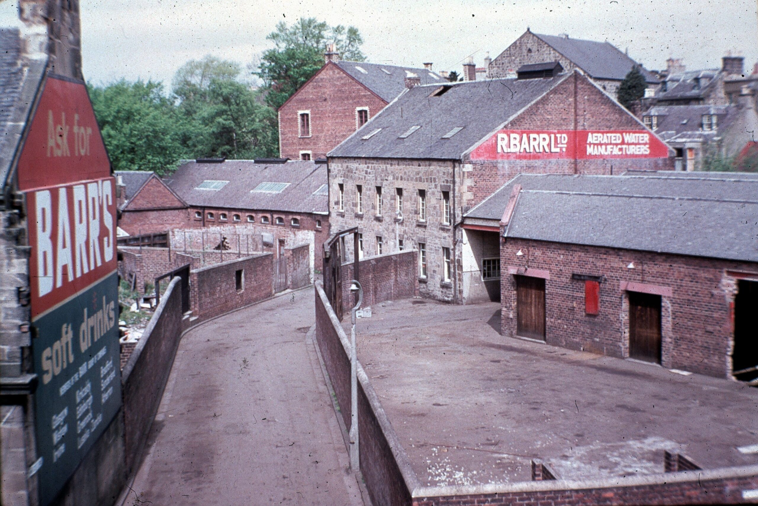 Colour photograph of the Barr's soft drinks factory complex, showing brick industrial buildings and a narrow lane leading through the site. Large painted Barr's advertising signs are visible on the buildings.
