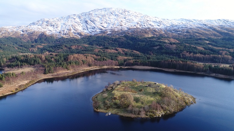 A small wooded island sits in a calm loch, surrounded by forested hills, with a snow-covered mountain rising in the background. The image is an aerial view taken on a clear day.
