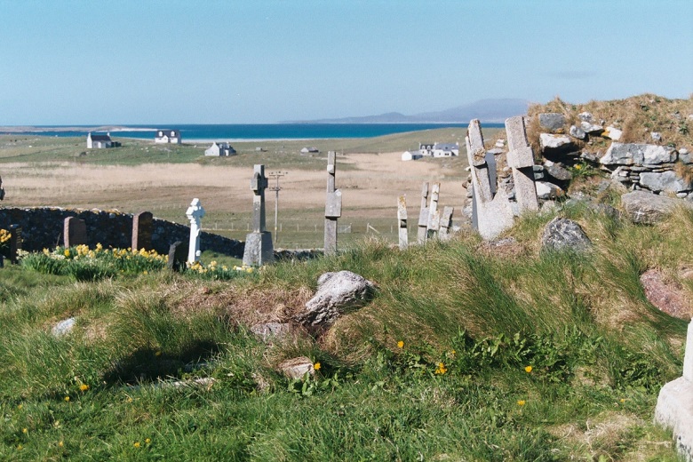 Weathered stone grave markers stand in a grassy cemetery beside a ruined stone wall, overlooking open fields and scattered white cottages. Beyond the settlement, a calm blue sea stretches towards distant hills.