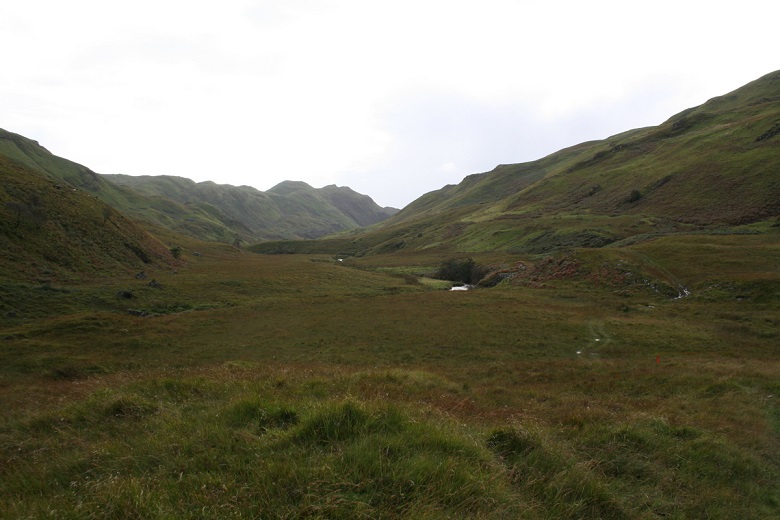 Wide view of a grassy glen surrounded by steep green hills. A small white building and a winding track sit near the centre of the landscape beneath an overcast sky.