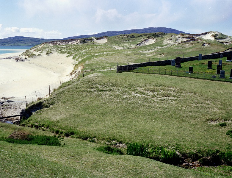 Grassy sand dunes and a sandy beach beside the sea, with a walled burial ground containing several gravestones on the right. Low hills rise in the background under a partly cloudy sky.