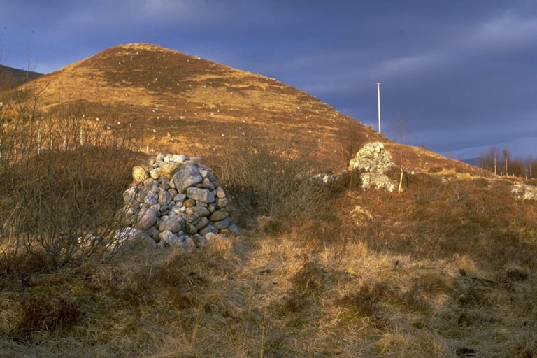 Two small cairns made of loose stones sit among scrub and grass at the foot of a rounded hillside. Low evening sunlight illuminates the brown slopes, while dark clouds fill the sky behind.