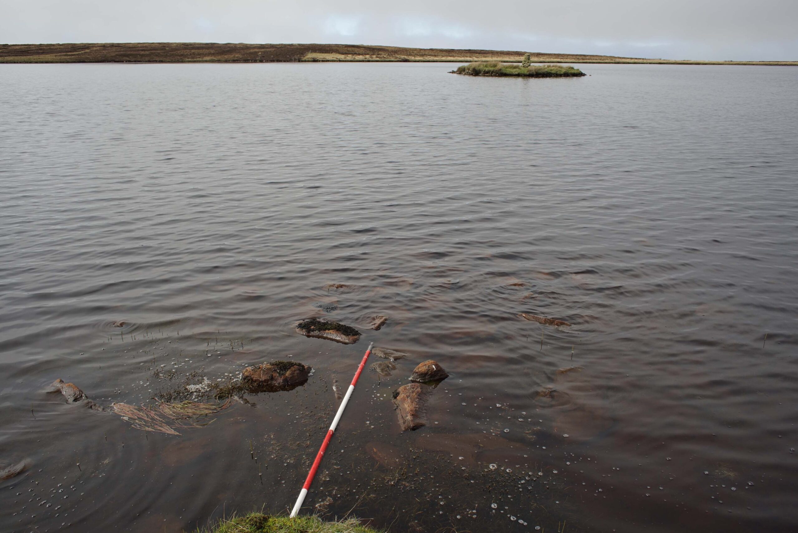 Small crannog island in a loch in the Western Isles, viewed from the shore with a line of stones leading towards the island.