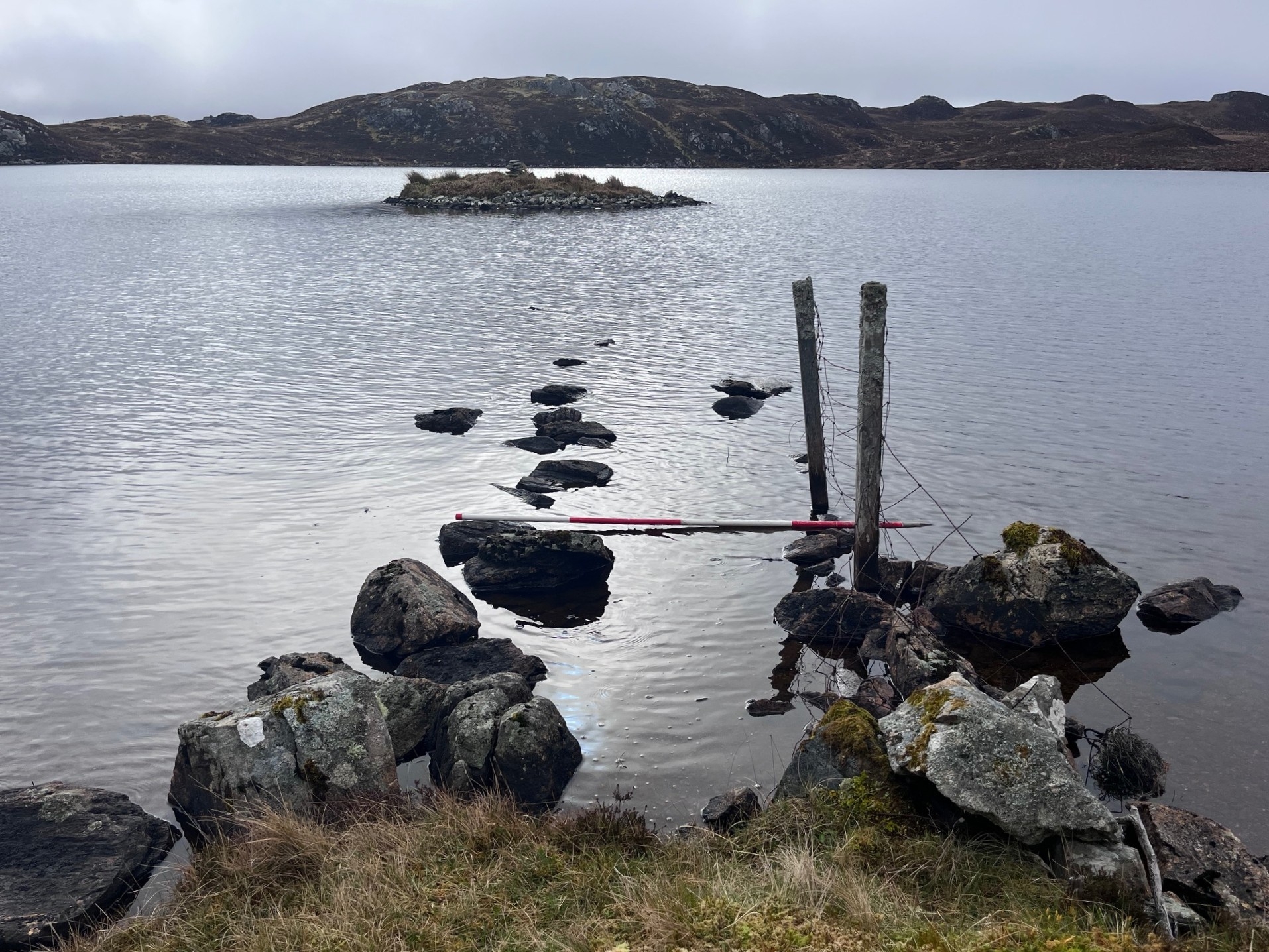 View across a loch towards a small crannog island in the Western Isles, with submerged stones visible in the foreground.