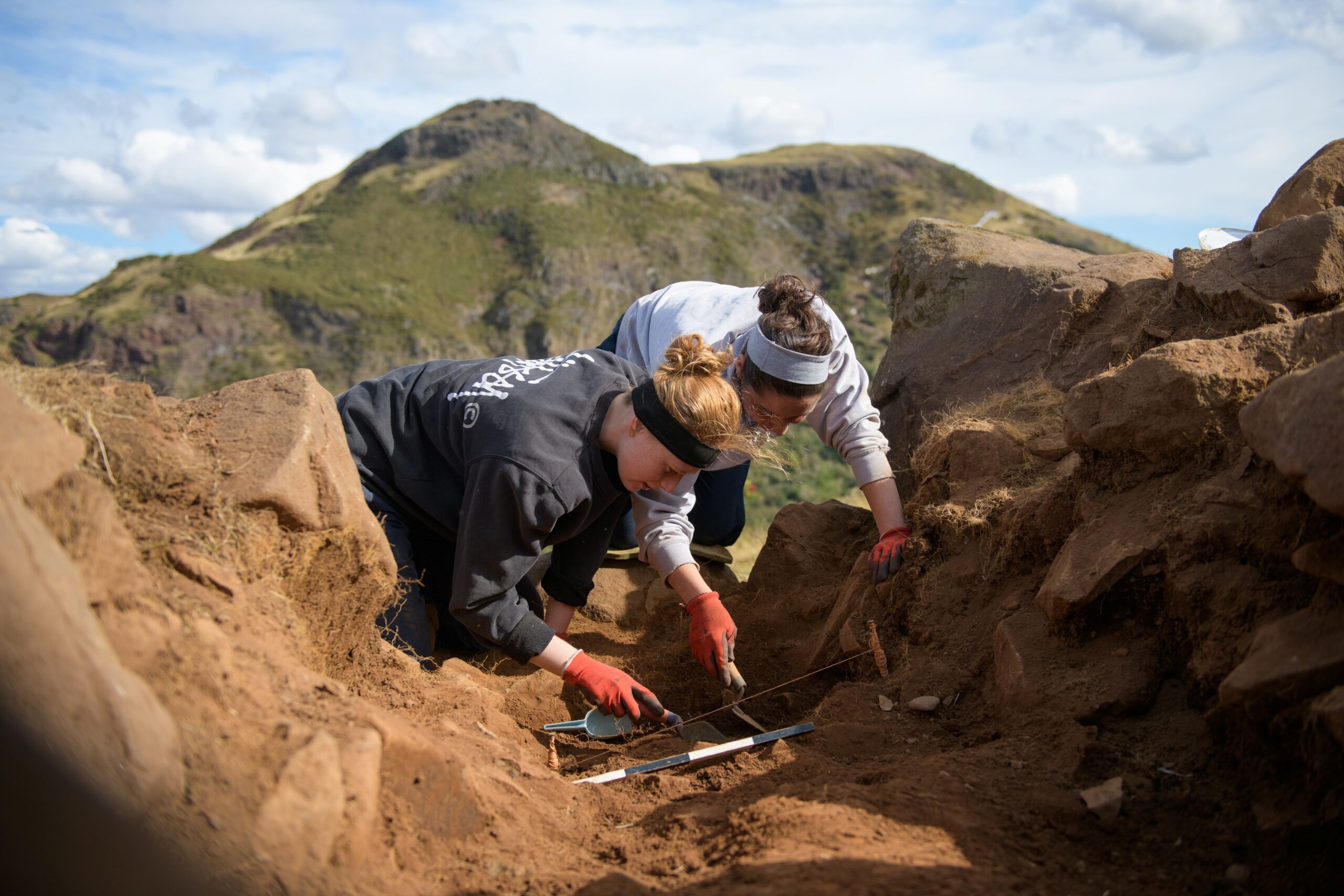 Two archaeologists working in dirt with hills and sky in the background. 