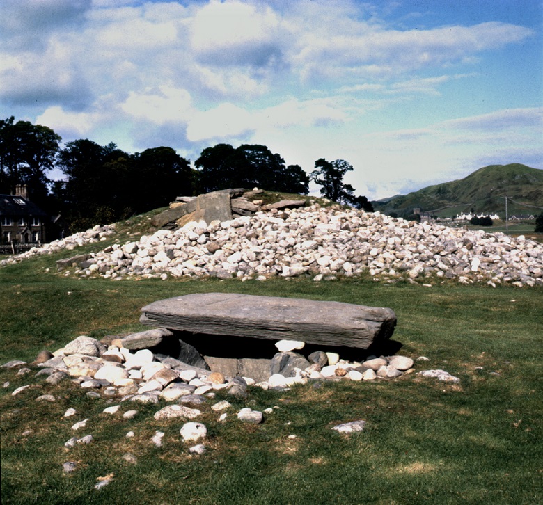 A large stone cairn under excavation stands in a grassy field, with a smaller stone-built feature in the foreground. Trees, houses and hills surround the site beneath a partly cloudy sky.