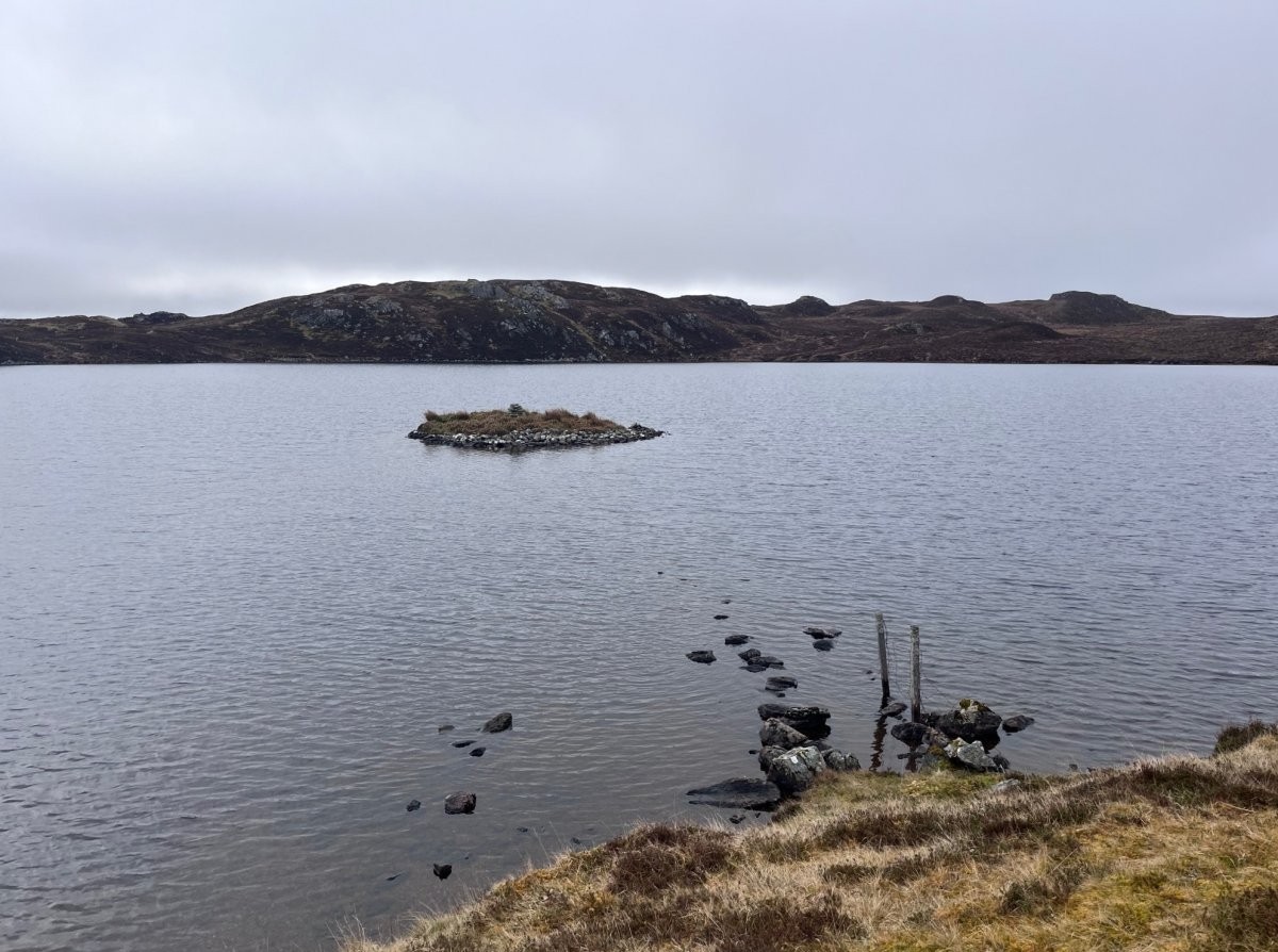 Small crannog island in the middle of a loch in the Western Isles, surrounded by open water under an overcast sky.
